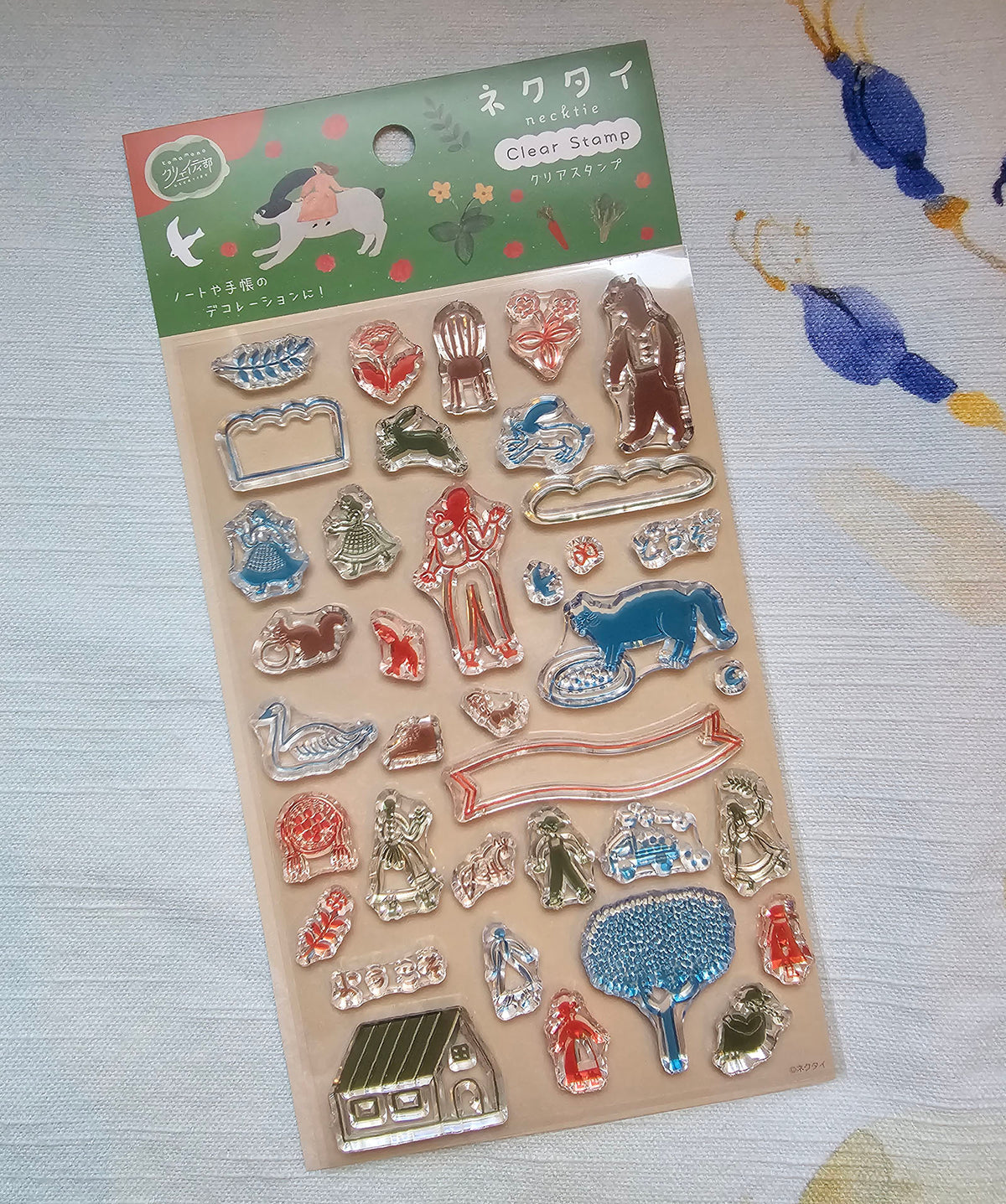 A package of Necktie clear stamps featuring various animal and decorative shapes in red, blue, green, and silver, with a green header card with Japanese text and illustrations, displayed on a patterned fabric background.