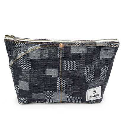 A rectangular patchwork pouch in a dark blue and gray denim-like fabric with a small copper-tone button, zipper closure, and a white brand patch reading 'Luddite' on the lower right. The pouch is photographed on a plain white background.