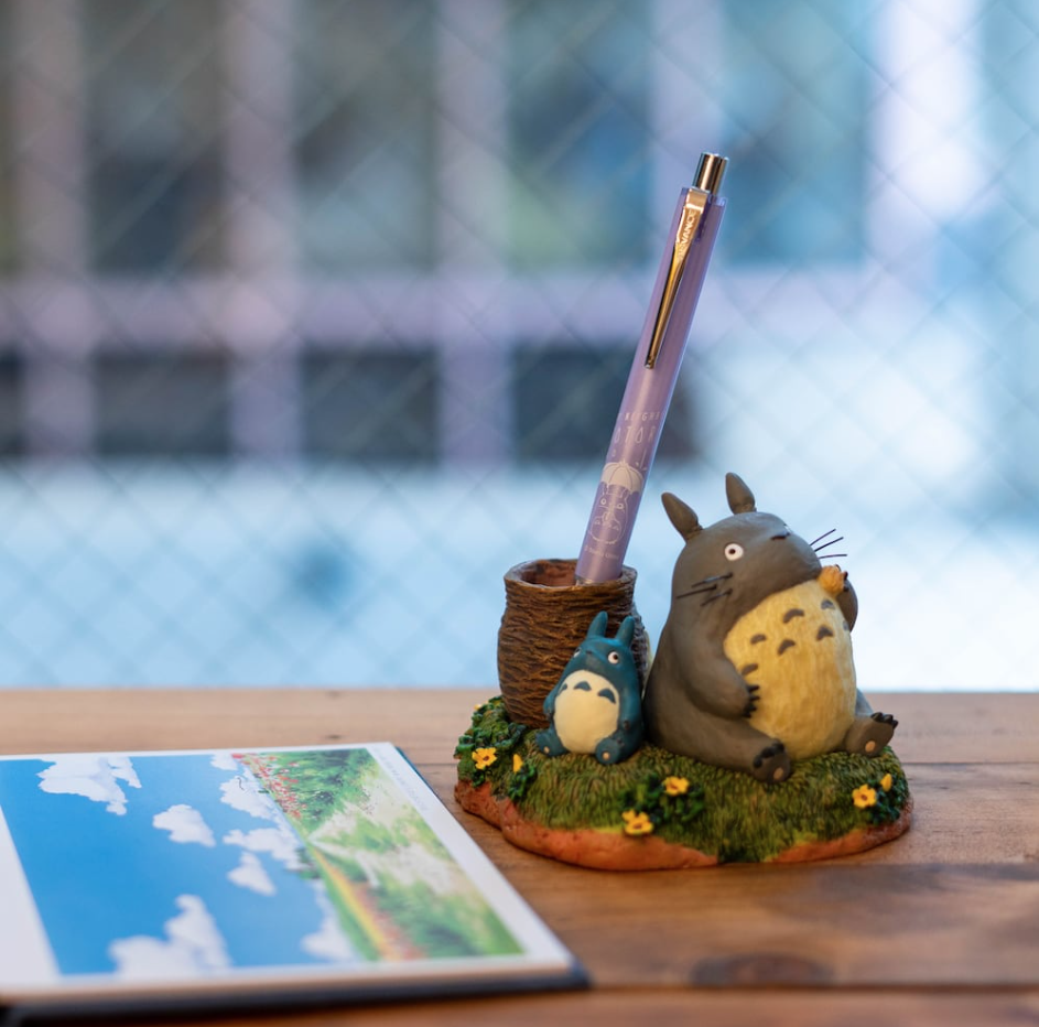 A Totoro-themed desk pen holder on a wooden tabletop, featuring a large gray Totoro figure holding a yellow acorn, a smaller blue Totoro figure, a mossy base with small flowers, a brown basket-like pen cup, and a lavender pen standing inside. A tablet screen showing a landscape is partially visible on the left, with a blurred window in the background.