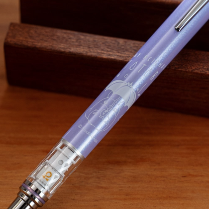A close-up of a translucent lavender pen resting diagonally on a wooden surface with a dark wood block in the background. The pen features a silver clip, a clear cap showing the ink reservoir, and white printed text and illustration of Totoro on the barrel.