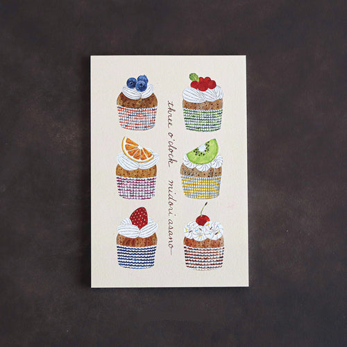 A postcard book featuring six illustrated cupcakes with different toppings, arranged on an off-white background with the phrase 'three o'clock' in script. The book is photographed flat against a dark textured background.