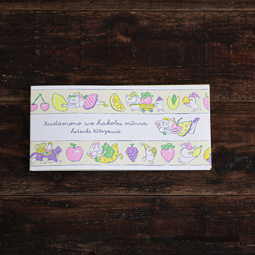 A rectangular printed card or notebook cover with a pastel illustrated border of fruits, flowers, and a cat, placed on a dark wooden surface. The center reads in Japanese: 'Kudamono wo hakobu minna'.