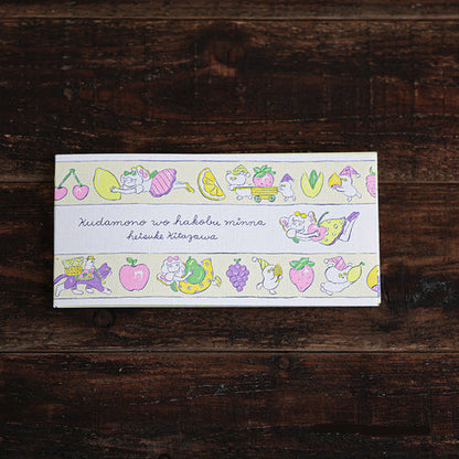A rectangular printed card or notebook cover with a pastel illustrated border of fruits, flowers, and a cat, placed on a dark wooden surface. The center reads in Japanese: 'Kudamono wo hakobu minna'.