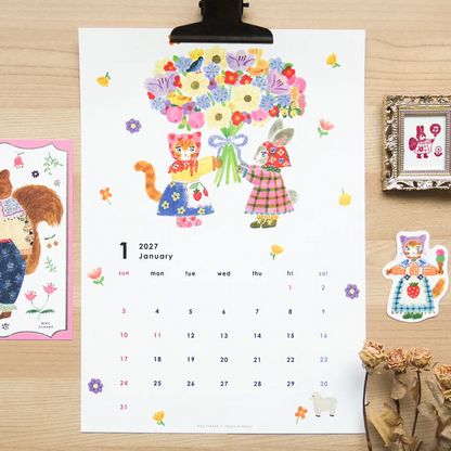 A printed January 2027 calendar page clipped at the top, featuring a whimsical illustration of two cats holding a large bouquet of flowers, displayed on a light wood surface with decorative stickers and framed art around it.