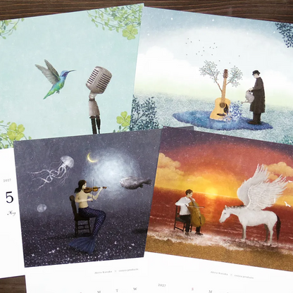Four illustrated calendar pages fanned out on a dark wooden surface. The designs show surreal scenes: a hummingbird and microphone, a tree and figure with a guitar, a mermaid playing violin under a crescent moon, and a white Pegasus in an orange sunset landscape.