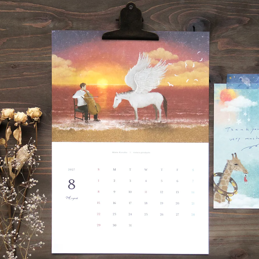 A printed desk calendar page clipped at the top, showing an illustrated scene of a seated figure beside a white winged horse over a sunset sky, with a calendar grid below. The page is photographed on a dark wood surface with dried flowers and a small card beside it.