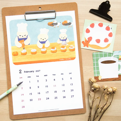 A wooden desk flat-lay featuring a monthly calendar printed on a brown clipboard, decorated with a cute illustration of polar bears making cupcakes. The clipboard is styled with a green pen, a black binder clip, a strawberry sticky note, a coffee mug note, and dried flowers on a light wood background.