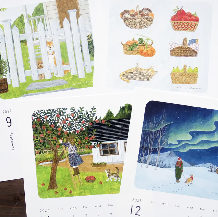 A close-up of a printed illustrated calendar pages showing multiple scenic panels: a cat behind a white picket fence, baskets of autumn produce, a snowy night scene with a person and dog, and monthly calendar dates visible along the left and bottom edges.