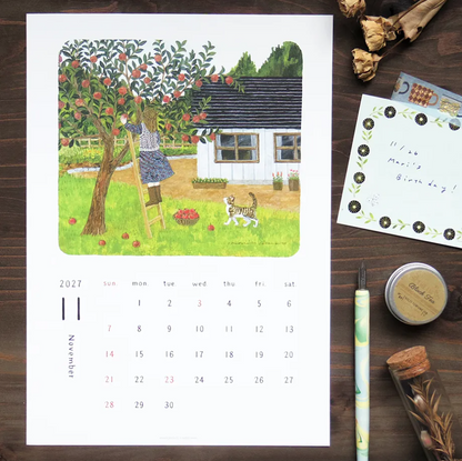 A printed monthly calendar page featuring a colorful watercolor-style illustration of a girl climbing a tree next to a small house, with a 2027 calendar grid below. The page is photographed on a dark wood surface with decorative stationery items around it, including a pen, dried flowers, a small tin, and a note card.