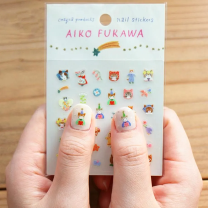 A person holds a package of Aiko Fukawa cat nail stickers. The clear plastic packaging shows various colorful cartoon cats and cat-themed designs, and the person's fingernails are decorated with two of the sticker designs.