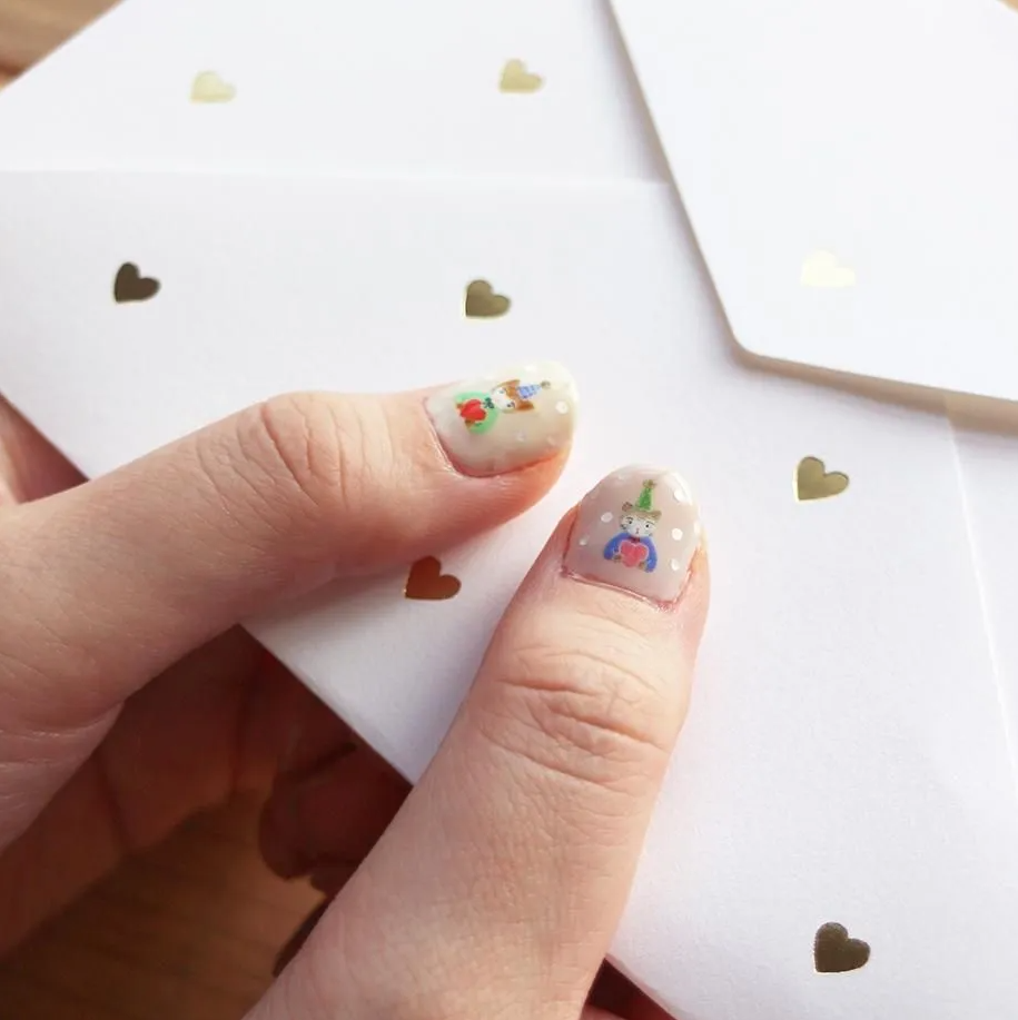A close-up of a hand holding white envelopes decorated with small metallic gold hearts. The fingernails are decorated with delicate, colorful nail art featuring cat characters.