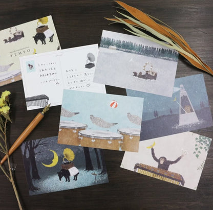 A flat-lay arrangement of illustrated postcards on a dark wood surface, alongside dried leaves, a wooden stick, and a small yellow flower. The postcards feature whimsical drawings of animals, music instruments, trees, and abstract scenes in soft pastel and muted tones.