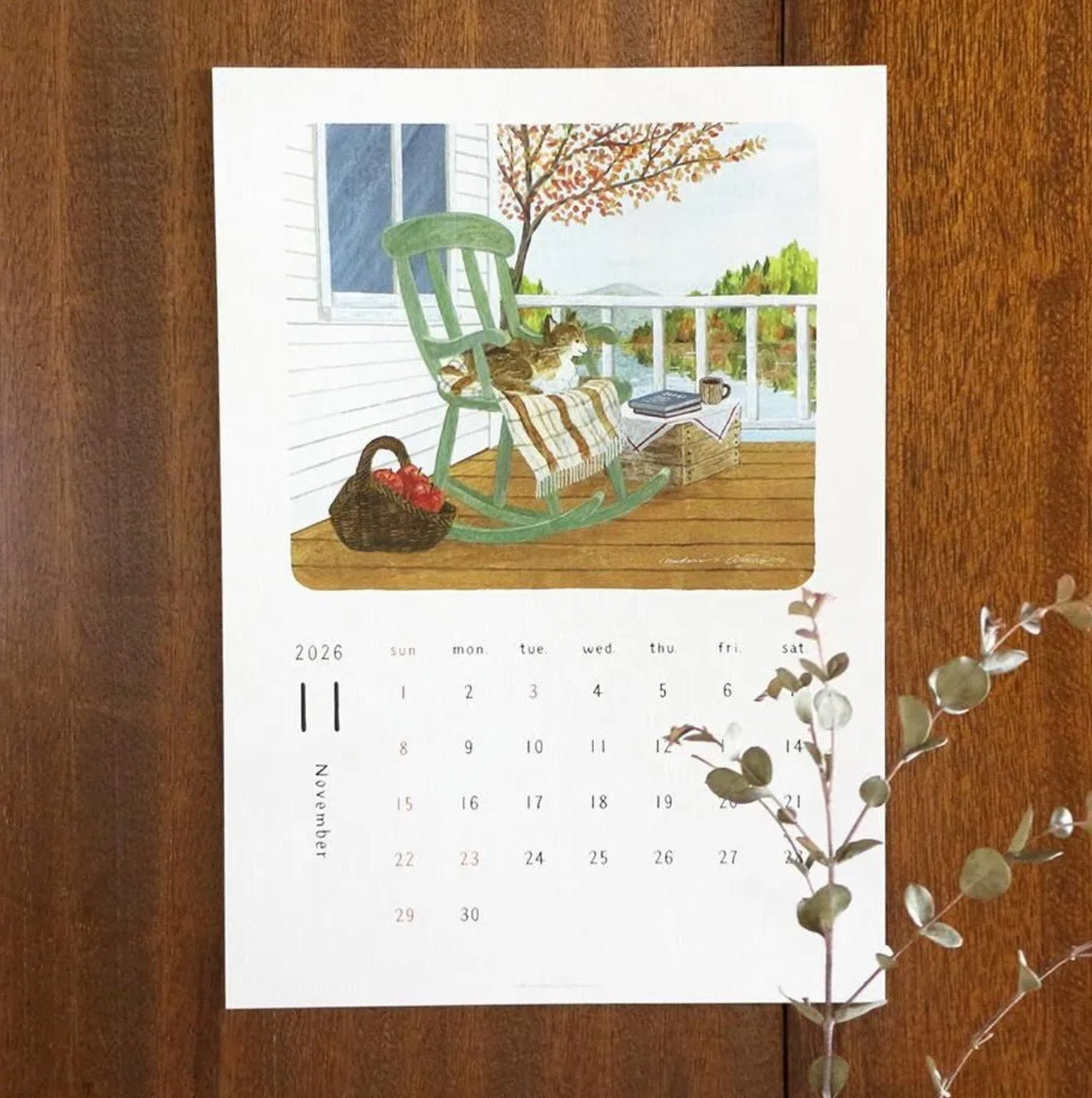 Calendar with illustration of a green rocking chair on a porch with a cat, displayed on a wooden surface.
