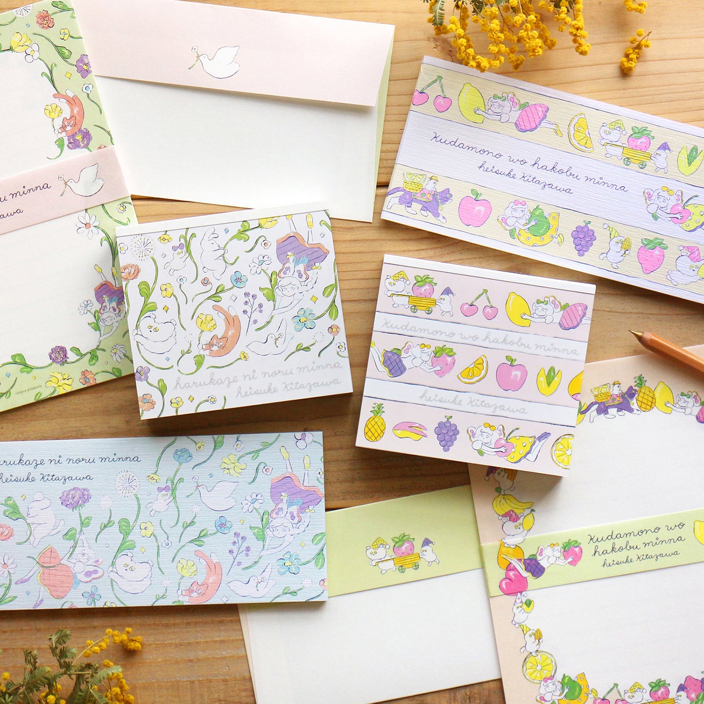 A flat-lay arrangement of cute pastel stationery items, including envelopes, cards, and decorative strips featuring cartoon animals and fruits, displayed on a light wood surface with small yellow flowers.
